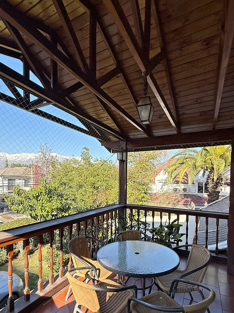 Balcón techado de madera con una mesa y sillas de mimbre; al fondo se ven árboles y la cordillera nevada