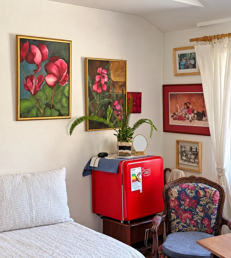 Habitación con cama de cobertor blanco, cuadros de flores en la pared, un frigobar rojo con un helecho encima, cortina blanca y un sillón de tapiz floreado