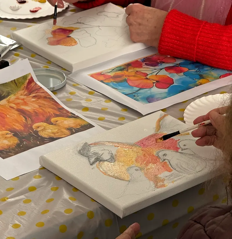Detalle del taller de pintura: una mano con pincel avanza sobre un lienzo de flores junto a láminas de referencia